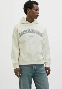 Felpa color crema con tasca a kangaroo frontale, grafica "JACK&JONES" in grigio scuro, polsini a coste e cappuccio con coulisse. Vestibilità casual.