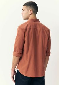 Chemise couleur rouille à manches longues, avec des poignets retroussés et un design au dos. Fabriquée en tissu lisse, elle dispose d'un ourlet droit et d'évasements latéraux.