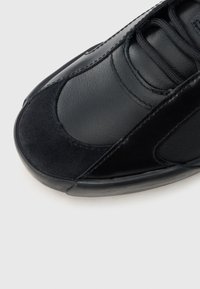 Črni športni čevlji z gladkim usnjenim zgornjim delom in semišnimi poudarki, zaobljenim sprednjim delom ter kontrastnimi teksturami. Zavezani z vezalkami in podrobnostmi šivov.