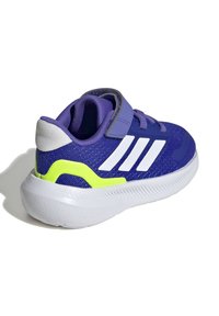 Zapatilla deportiva morada con acentos blancos, parte superior texturizada y tobillo acolchado. Presenta una correa de Velcro y una suela blanca gruesa con detalles verdes.