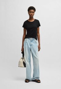Maglietta nera a maniche corte, jeans azzurri larghi, sandali neri, che tiene in mano un secchiello beige e nero. Design minimalista con texture morbide.