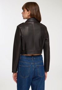 Młoda kobieta w krótkiej czarnej skórzanej kurtce i niebieskich dżinsach o wysokim stanie, stojąca tyłem na jednolitym jasnym tle.