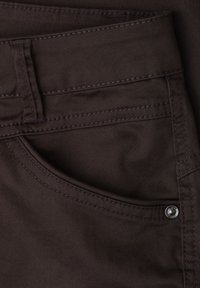 Gros plan sur un tissu marron foncé montrant une poche avant style jean, une boucle de ceinture et un détail de rivet métallique sur le côté droit.