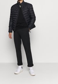 Schwarze gesteppte Jacke mit hohem Kragen, kombiniert mit einem schwarzen Pullover, hochgekrempelten schwarzen Hosen und weißen Turnschuhen. Durchgehend glatte Textur.