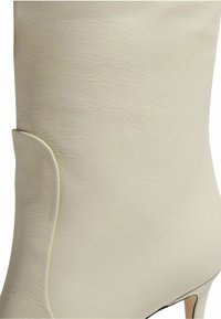 Bota de tacón alto de cuero crema con una textura suave y elegante y una sutil curvatura en el tobillo, sin herrajes visibles.