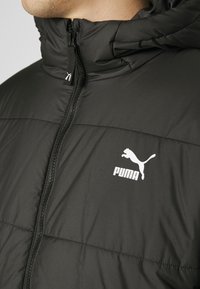 Černá péřová bunda s vysokým límcem, zipovým zapínáním a bílým logem Puma na levé hrudi. Materiál je hladký a texturovaný.