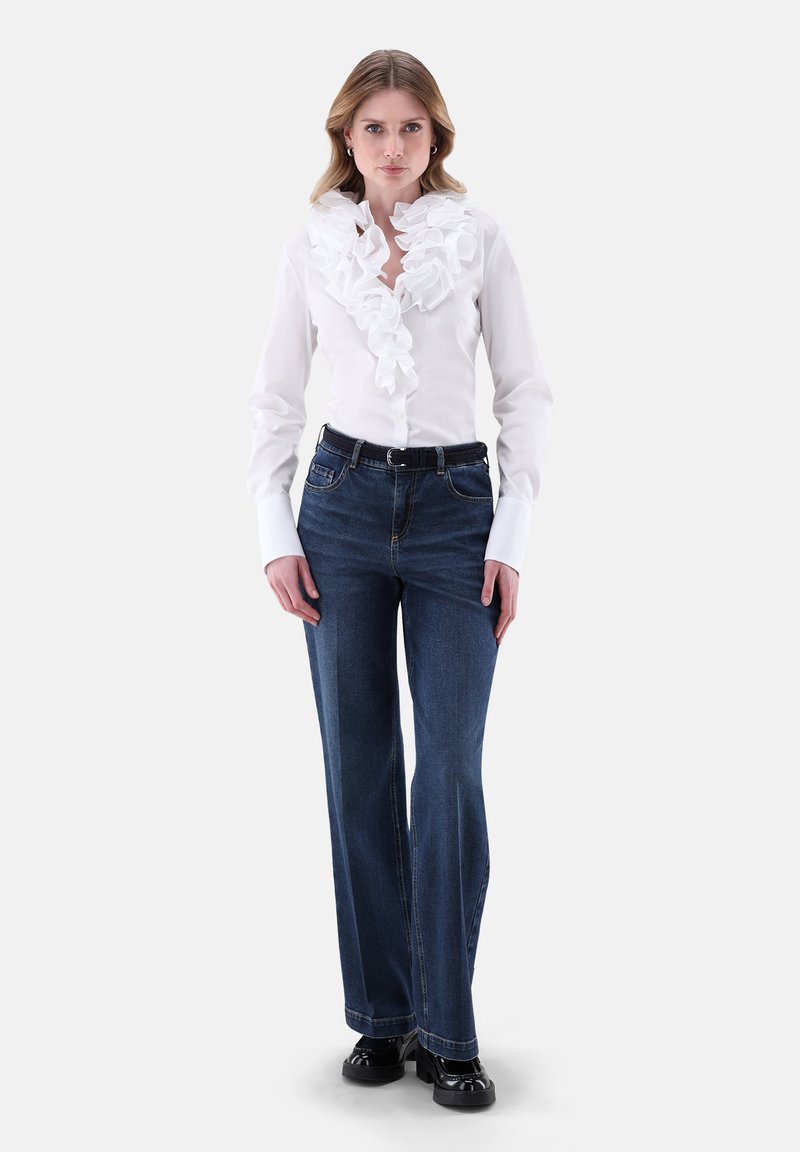 Weißes Hemd mit Rüschenschnitt, kombiniert mit dunkelblauen Jeans im Schlagstil. Schwarzer Gürtel und Schuhe runden das Outfit ab.