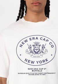 Hvid bomulds t-shirt med et marineblåt grafisk tryk indeholdende "New Era Cap Co" og et våbenskjold-design, herunder tekst om kvalitet og etableringsår.