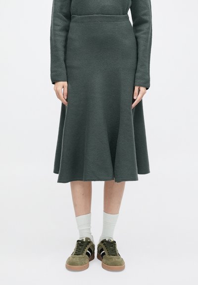 Σκούρο πράσινο, φούστα με φαρδιά γραμμή, φτιαγμένη από υφασματισμένο ύφασμα, συνδυασμένη με ελαιόχρωμα αθλητικά παπούτσια και ανοιχτόχρωμες κάλτσες. Απλός σχεδιασμός, μήκους μέχρι το γόνατο.