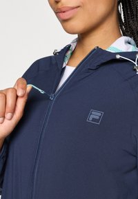 Navy zip-up jacket with hood, featuring a toggle and interior patterned lining. Displaying a logo on the chest and smooth texture.