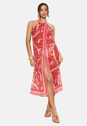 Naine punase ja roosakirju halterkaelusega kleidis, millel on ees lõhik, kandmas kuldseid käevõrusid ja pruune sandaale, seisab tavalise tausta ees.