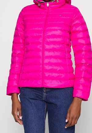 Femme portant une veste matelassée rose vif avec fermeture éclair, capuche, et jean bleu moyen, debout devant un fond clair.