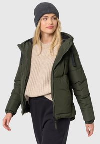 Olijfgroene pufferjack met rits, capuchon en elastische manchetten, gedragen over een beige gebreide trui, gecombineerd met zwarte sweatpants en een grijze beanie.