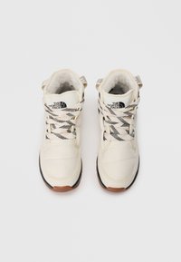 Light cream high-top boots with black rubber soles. Features textured fabric, thick black-and-white laces, and a branded logo on the side.