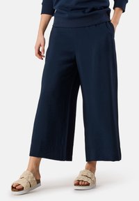 Marineblau weite Hosen aus einem weichen, strukturierten Stoff. Kombiniert mit geflochtenen beigen Sandalen, die eine weiße Sohle und strukturierte Riemen haben.