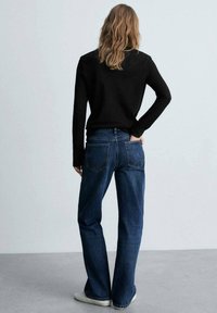 Maglietta nera a maniche lunghe abbinata a jeans blu affusolati con due tasche posteriori e un fit rilassato; il modello si trova in posa con le mani sui fianchi.