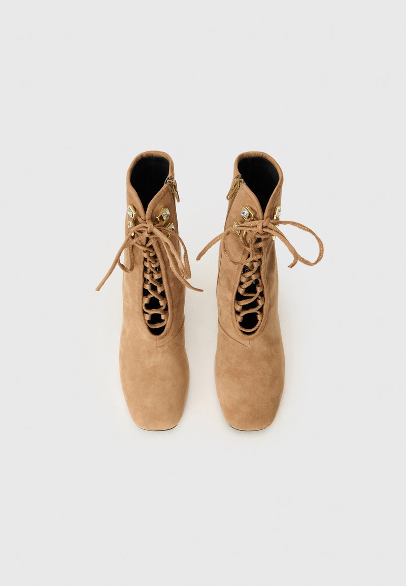 Beige Wildleder-Ankle Boots mit einem ausgeschnittenen Schnürdesign, das goldene Hardware-Details und eine glatte Textur aufweist. Quadratische Zehen und ein schlanker Absatz.