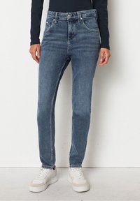 Svetle modré džíny s vysokým pasem vyrobené z denimu. Klasický střih s pěti kapsami. Spárovány s bílými teniskami a tmavým topem s dlouhým rukávem.