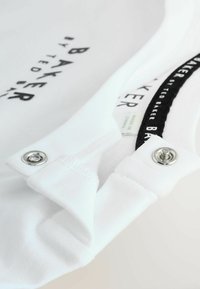 Indumento in tessuto bianco con colletto a bottoni, applicazioni in metallo e un'etichetta stampata in bianco e nero, con texture rigata visibile.
