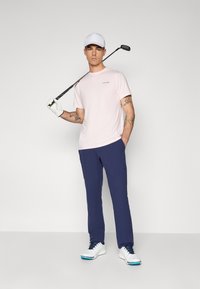 Roza majica s kratkimi rokavi z logotipom, mornarske golf hlače in bele golf čevlje z modrimi detajli. Drži golf palico in nosi belo kapo.