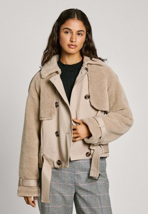 Veste cropped beige avec une texture moelleuse, un design double boutonnage, de grands boutons et une taille ceinturée. Le modèle porte un pull noir en dessous.