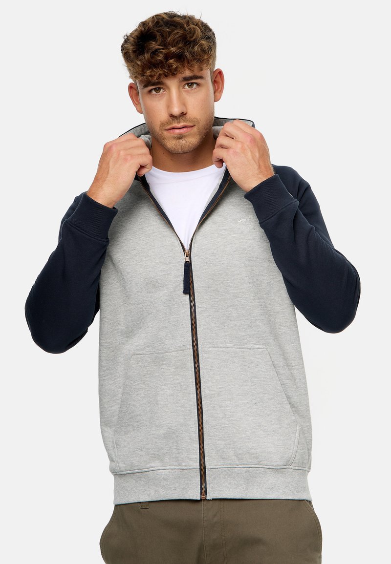 Grijze zip-up hoodie met marineblauwe mouwen, gemaakt van een katoenmix. Beschikt over een opstaande kraag en een kangoeroe-pocket. Bruine ritsdetails.