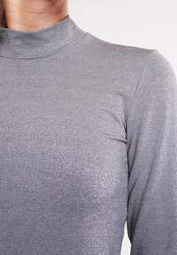 Harmaa metallinen pitkähihainen paita, jossa on korkea kaulus ja sileä, säihkeä pinta. Kankaan pinta heijastaa valoa, mikä korostaa muotoilua.