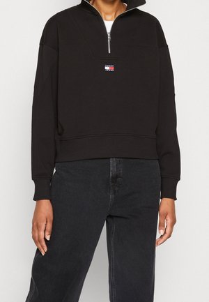 Person iført en sort quarter-zip sweatshirt med Tommy Jeans-logo og højtaljede sorte denimjeans mod en ensfarvet baggrund.