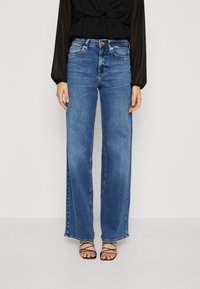 Weite blaue Jeans mit hellem Washedesign, ausgestattet mit einem klassischen Fünf-Taschen-Schnitt und einem Knopfverschluss an der Taille. Schwarze Riemchensandalen.