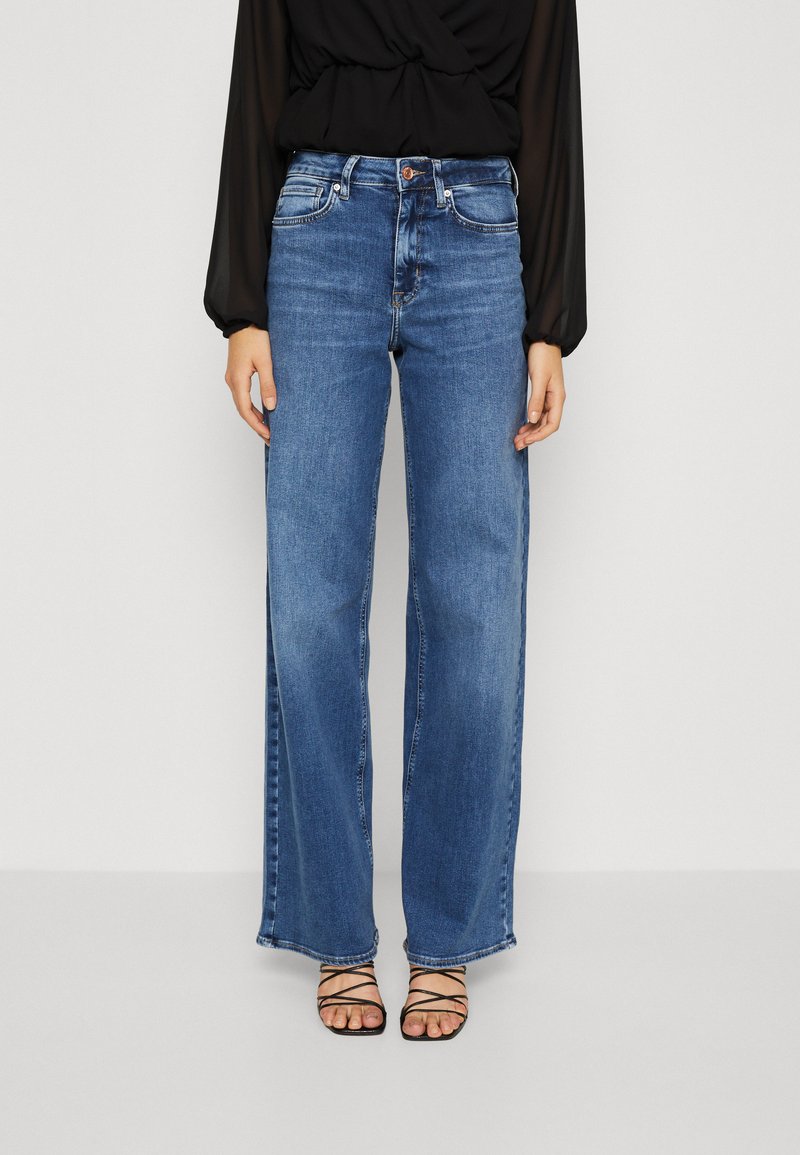 Weite blaue Jeans mit hellem Washedesign, ausgestattet mit einem klassischen Fünf-Taschen-Schnitt und einem Knopfverschluss an der Taille. Schwarze Riemchensandalen.
