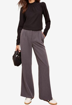 Femme portant un haut noir à manches longues et un pantalon gris à jambes larges, tenant un sac à main noir tissé, debout avec une main dans la poche.