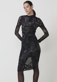 Rochie neagră din dantelă florală, cu mâneci lungi, siluetă ajustată și un overlay transparent. Prezintă o fente până la genunchi și detalii din țesătură texturată.