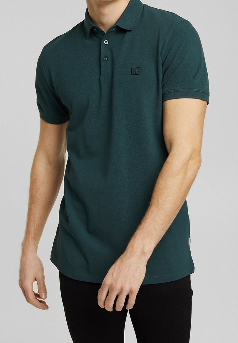 Homme portant un polo vert foncé à manches courtes avec un petit logo sur la poitrine gauche, associé à un pantalon noir.