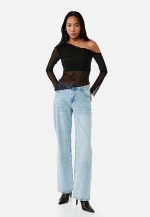 Kobieta w czarnym, przezroczystym topie na jedno ramię, jasnoniebieskich, prostych wysokich dżinsach i czarnych botkach na obcasie ze spiczastym noskiem, stojąca z rękami w kieszeniach.