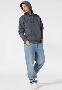 Grå hættetrøje med forlomme og broderet logo, kombineret med lyseblå løstsiddende jeans og hvide sneakers.