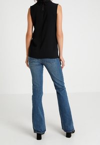 Sort ærmeløs bluse med høj krave og nøglehulsryg, parret med blå flagrende jeans og sorte hælskotter. Glat tekstur, afslappet stil.