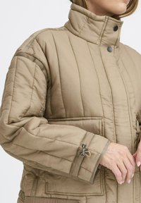Beige quiltet jakke med høj krave, trykknapper og sidelommer. Har en struktureret overflade og justerbar manchette med hardware detaljer.