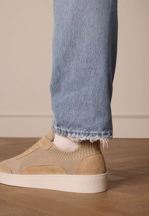 Baskets beiges en mesh texturé et en daim, avec une semelle blanche. Portées avec un jean déchiré bleu clair et des chaussettes blanches.