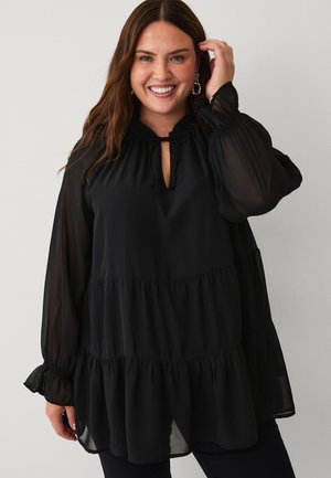 Smilende kvinde med langt brunt hår iført øreringe og en løs, lagdelt sort bluse med gennemsigtige lange ærmer, stående mod en ensfarvet baggrund.