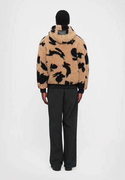Man in een beige en zwart gestreepte sherpa-jas met capuchon en zwarte broek, staand met zijn rug naar de camera tegen een effen witte achtergrond.