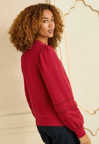 Červený pulóver so zúženými rukávmi, s čipkovými detailmi na manžetách. Vyrobený z mäkkej tkaniny, navrhnutý pre voľný strih.