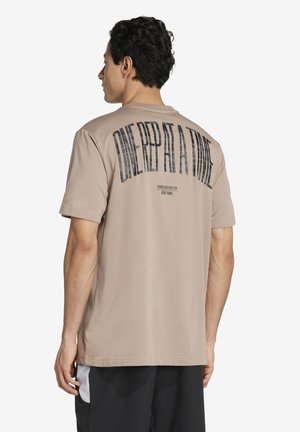 Mann, der mit dem Rücken zur Kamera steht, trägt ein beiges T-Shirt mit dem großen Schriftzug "ONE REP AT A TIME" auf dem Rücken und schwarze Shorts mit weißen Akzenten.