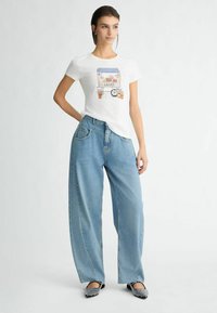 T-shirt en coton blanc avec un imprimé graphique coloré représentant un stand de marché. Assortie à un jean en denim large de couleur bleu clair et des ballerines noires.