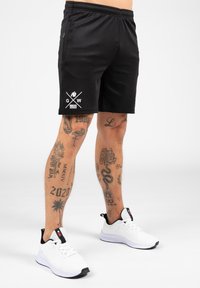 Zwarte sportshorts met een elastische tailleband, voorzien van een wit logo en gekruiste lijnen op de linker dij. Geselecteerd met witte sneakers.