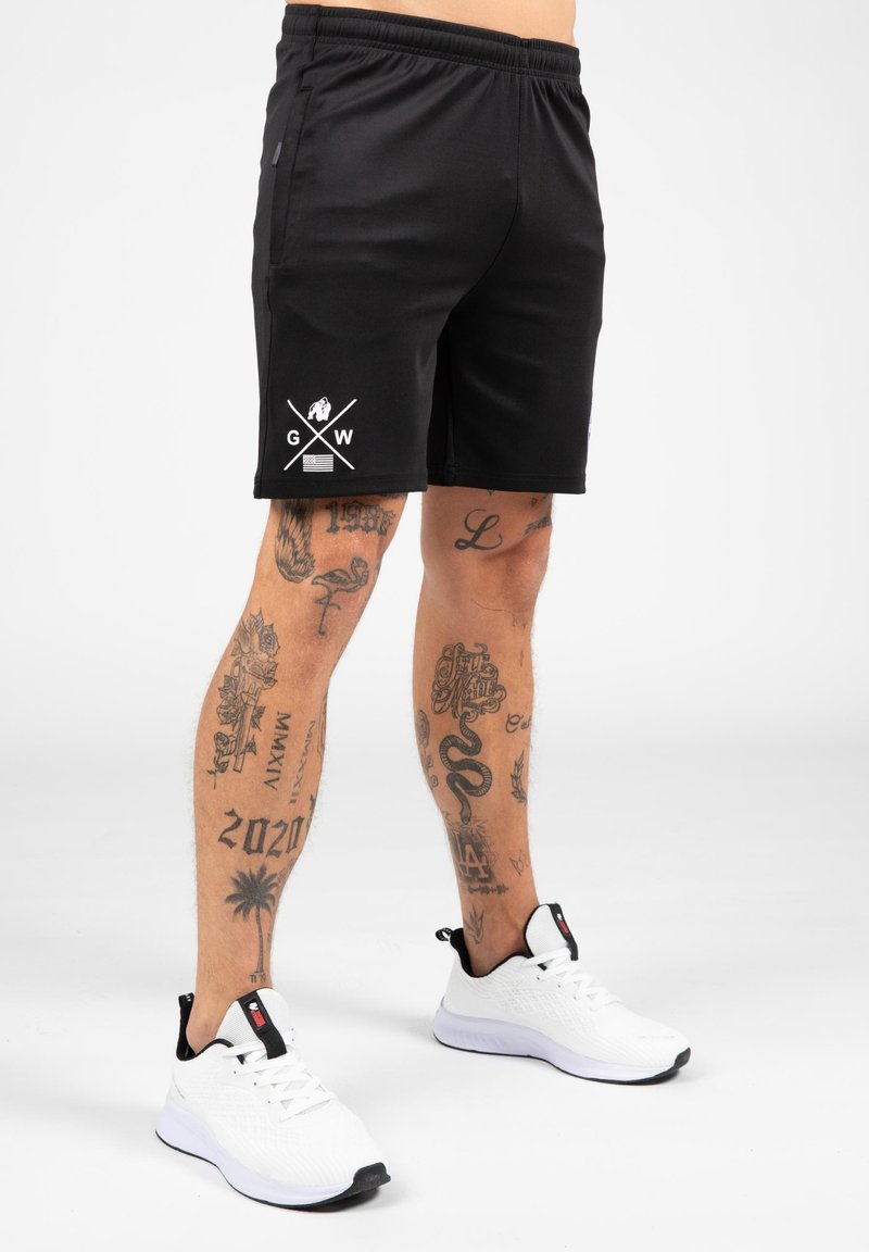 Zwarte sportshorts met een elastische tailleband, voorzien van een wit logo en gekruiste lijnen op de linker dij. Geselecteerd met witte sneakers.