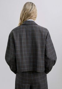 Blazer recortado en tela gris oscuro con un patrón de cuadros que presenta acentos azules. Diseñado con cuello de muesca y corte relajado.