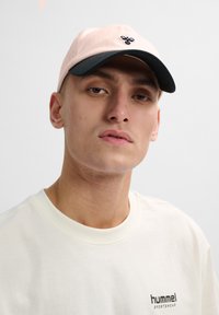 Light pink baseball cap with a dark brim, featuring a small black logo. Paired with a cream-colored short-sleeve shirt.