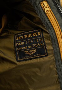 Czarny etykieta na oliwkowym tkaninie z żółtymi akcentami. Tekst zawiera "SKYTRUCKER", numer wydania oraz markę "PME LEGEND." Widoczny zamek.