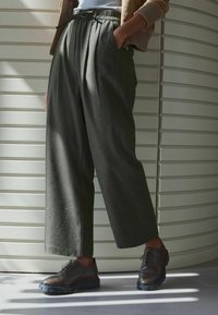 Pantalons plissés vert foncé avec taille à cordon, associés à des chaussures en cuir marron avec des détails en brogue et une épaisse semelle en caoutchouc.