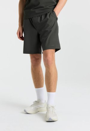 Mand iført sorte shorts, hvide sokker og hvide sportssko, der står med den ene hånd i lommen på en ensartet lys baggrund.
